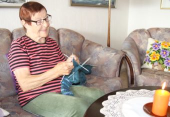 Iloinen vanhempi nainen neuloo sohvalla, pöydällä palaa kynttilä.