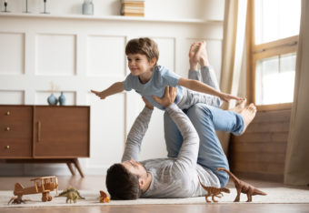 Poika leikkii lentokonetta olohuoneen lattialla makaavan isän käsien ja jalkojen päällä.