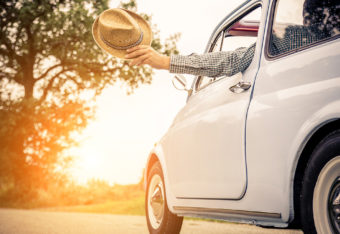 Kesäauto, auton ikkunasta ojennetussa kädessä aurinkohattu.