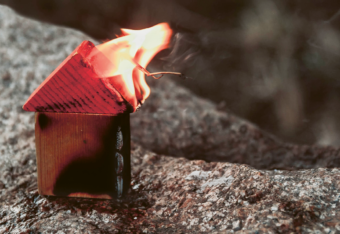 Kallion päällä liekehtivä puupalikoista tehty pikkutalo.