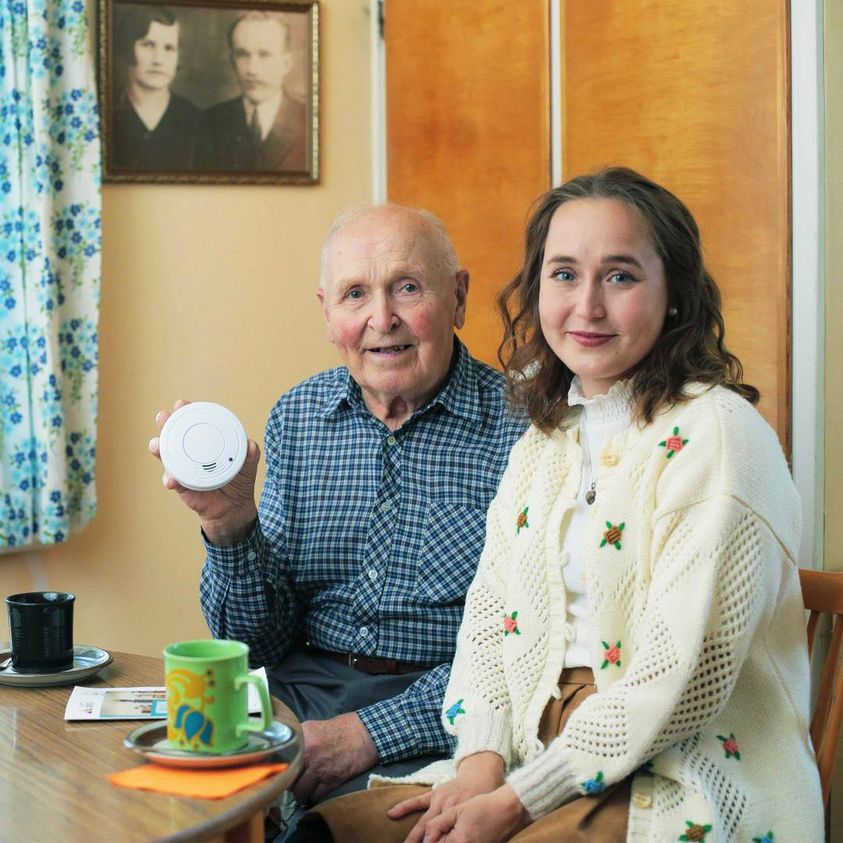 Sosiaalisen median kasvot Lotta ja Pappa kahvilla, ja Pappa näyttää kädessään olevaa palovaroitinta.