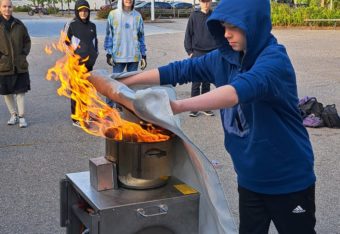 Yläkouluikäinen poika harjoittelee sammuttamaan kattilassa olevaa tulipaloa sammutuspeitteellä.