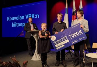 Kolme Kouvolan VPK:n palokuntanuoriso-osaston lasta seisoo lavalla iso rahasekki käsissään.