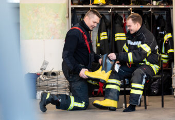 Palokuntalainen auttaa uutta palokuntalaista sovittamaan varusteita.