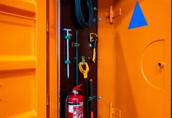 Oranssi suojakontti, johon on maalattu sininen kolmio. Kontissa on pelastajien tarvikkeita kuten lapio, kirves ja käsisammutin.