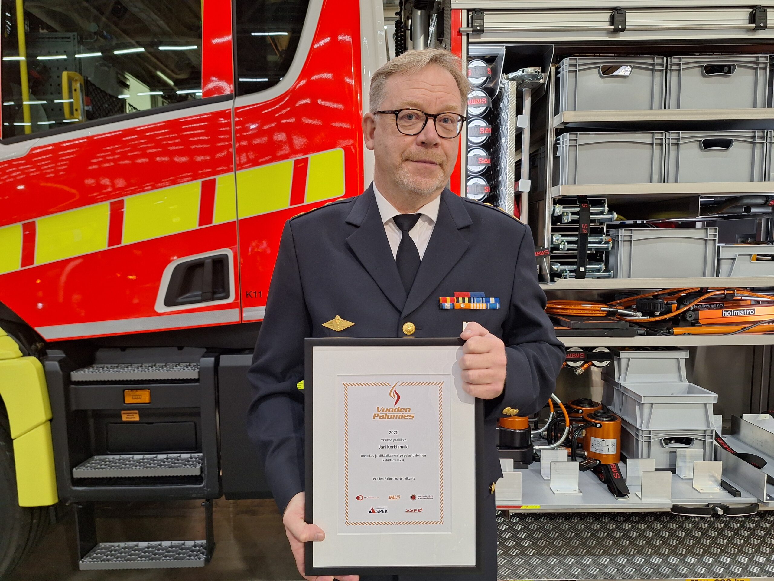 Vuoden palomieheksi valittu mies seisoo palkintotaulu kädessään paloauton edessä.
