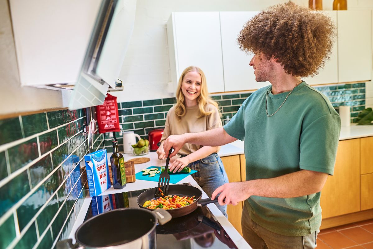Kaksi nuorta aikuista laittaa iloisina ruokaa keittiössä lieden ääressä.