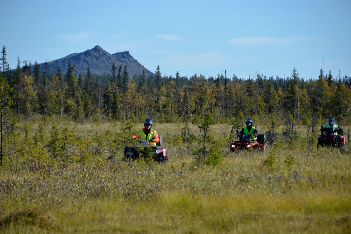 Kolme ihmistä ajaa mönkijöillä metsäisessä erämaassa, jonka taustalla kohoaa vuori.