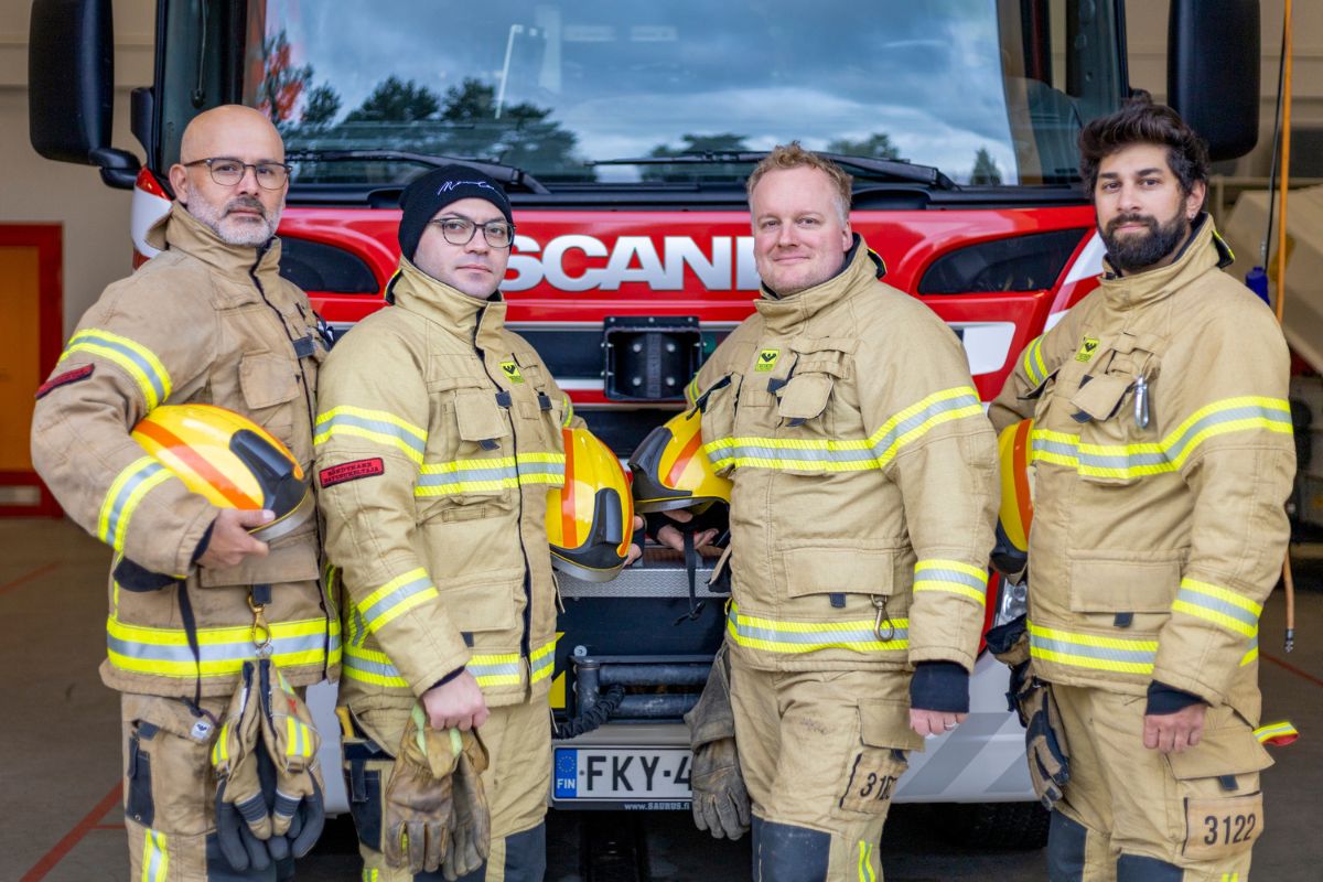 Neljä sopimuspelastajan varusteisiin pukeutunutta miestä seisoo paloauton edessä ja pitää käsissään kypäröitä.