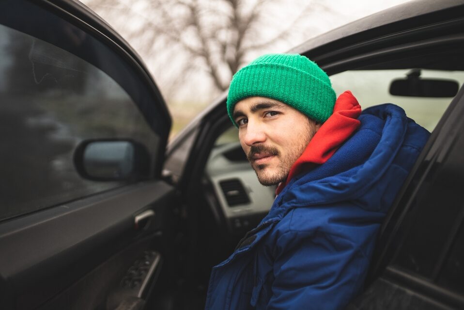 A young man with a beanie and a mustache sits in a car behind the wheel, the door open, looking toward the camera with a mysterious expression.