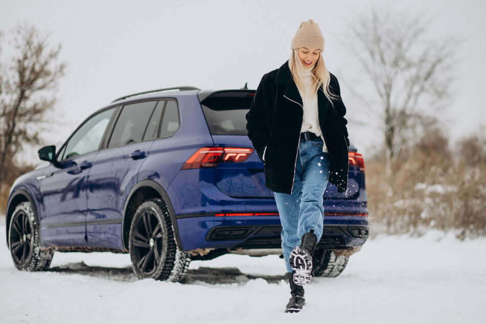 A smiling young woman wearing a beanie kicks snow in front of her. A blue car is in the background.