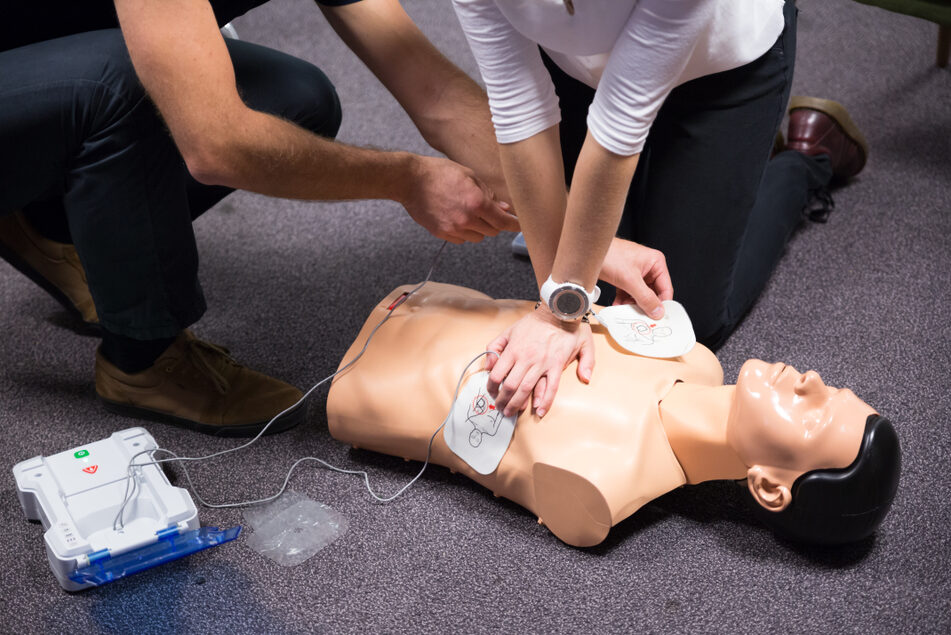 A CPR training mannequin with defibrillator pads being placed on its chest. At the same time another person is performing chest compressions.