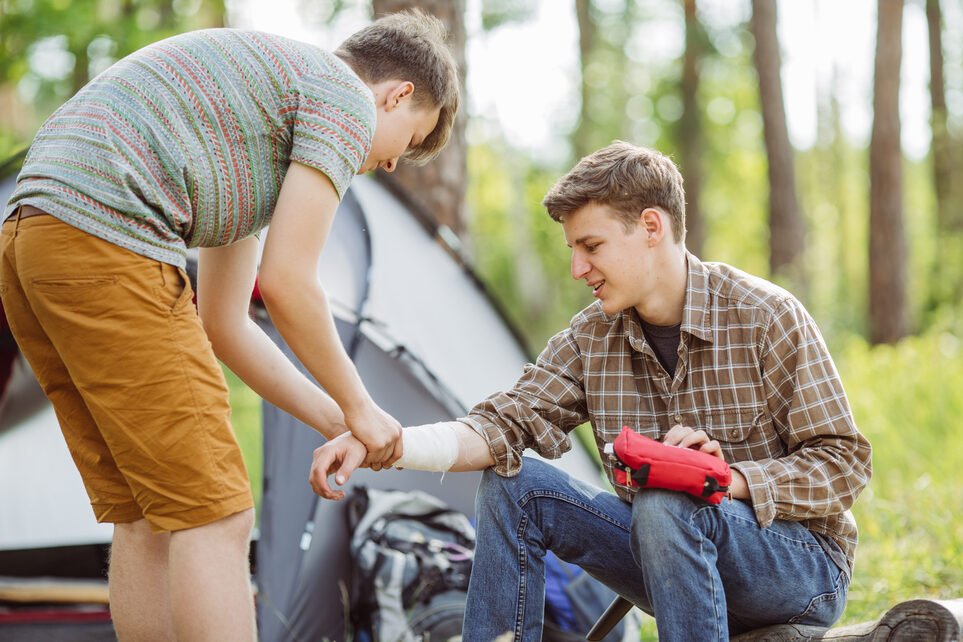A person dressed in outdoor clothing sits on the ground in the forest, holding one ankle with both hands.