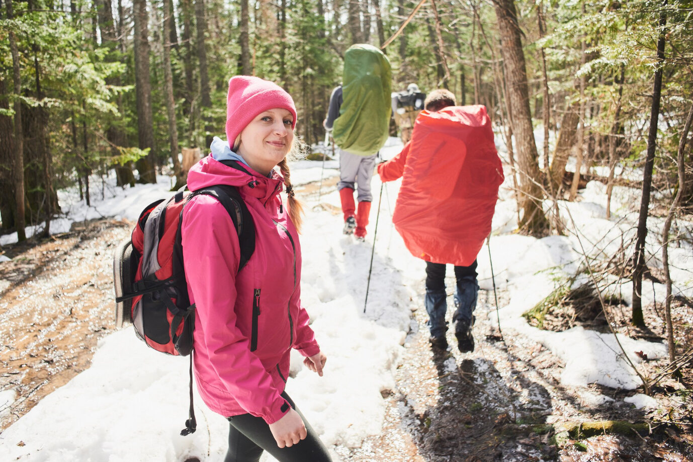 A small group of people hikes through a snowy forest in sunny weather. They carry backpacks and use walking poles.