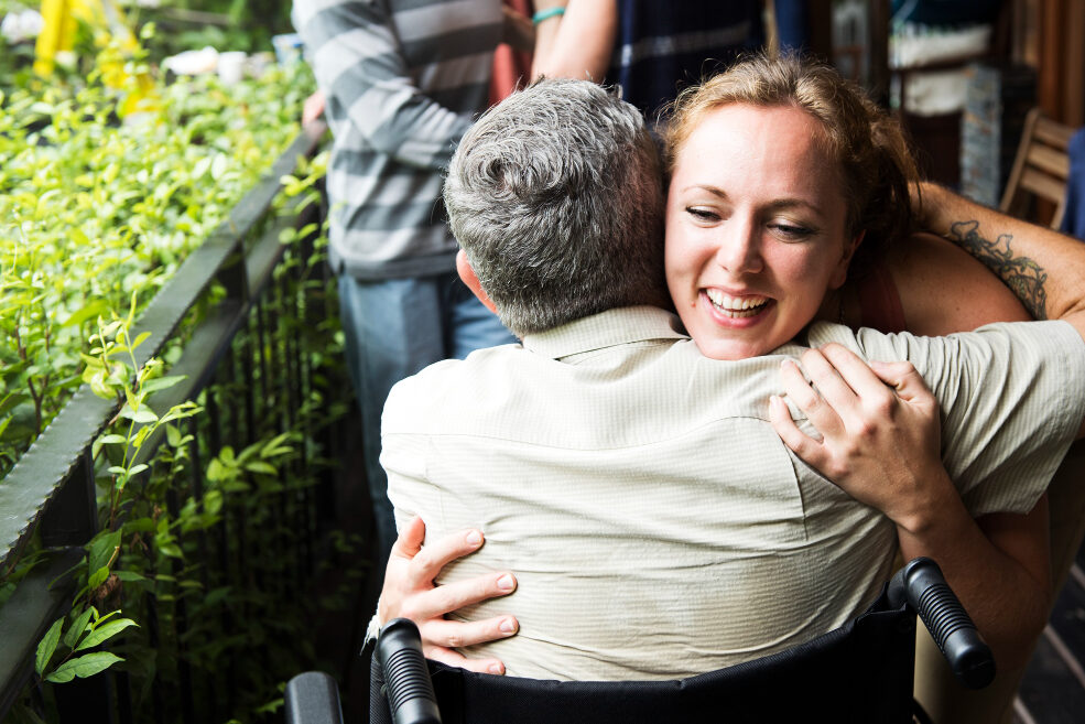 A smiling woman hugs an older person in a wheelchair on an outdoor terrace.