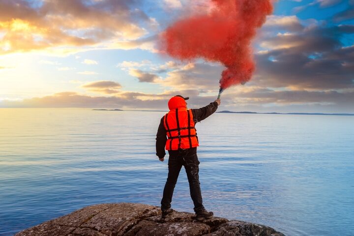 A person in a life jacket stands on a high cliff, waving a red distress flare in one hand.