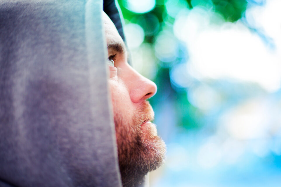 A bearded young man wearing a hood looks thoughtfully upward.