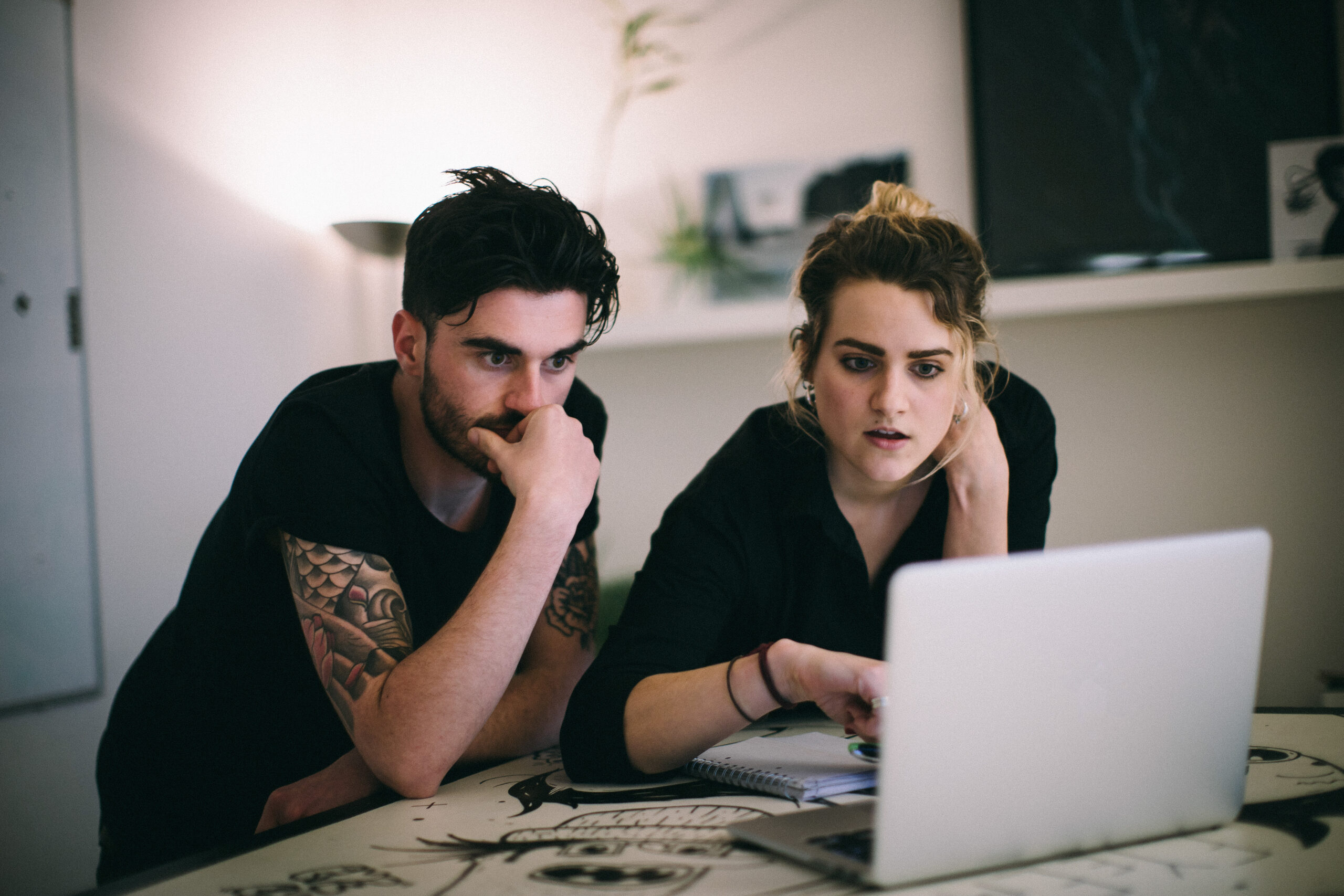 A serious-looking young woman scrolls on a laptop while a young man next to her rests his hand over his mouth, also staring seriously at the screen.