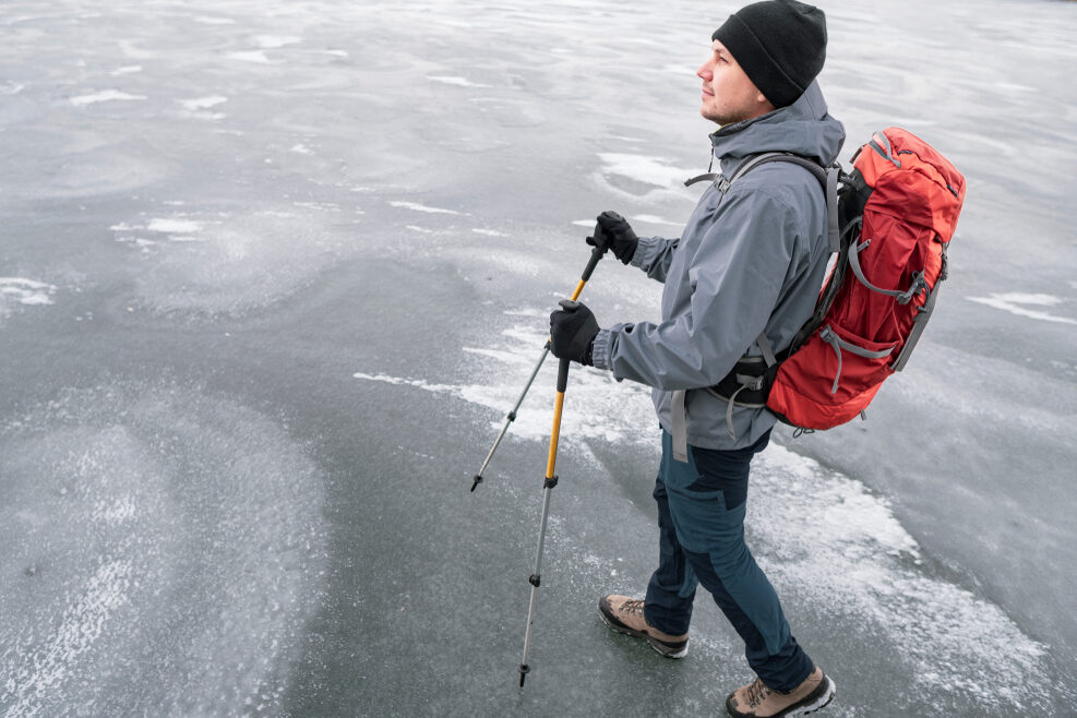 A young adult walks on a frozen lake or sea, holding walking poles and carrying a small backpack.