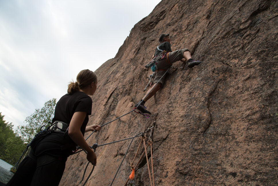 Two people rock climbing in Repovesi National Park. One is climbing while the other belays from below, holding the rope.