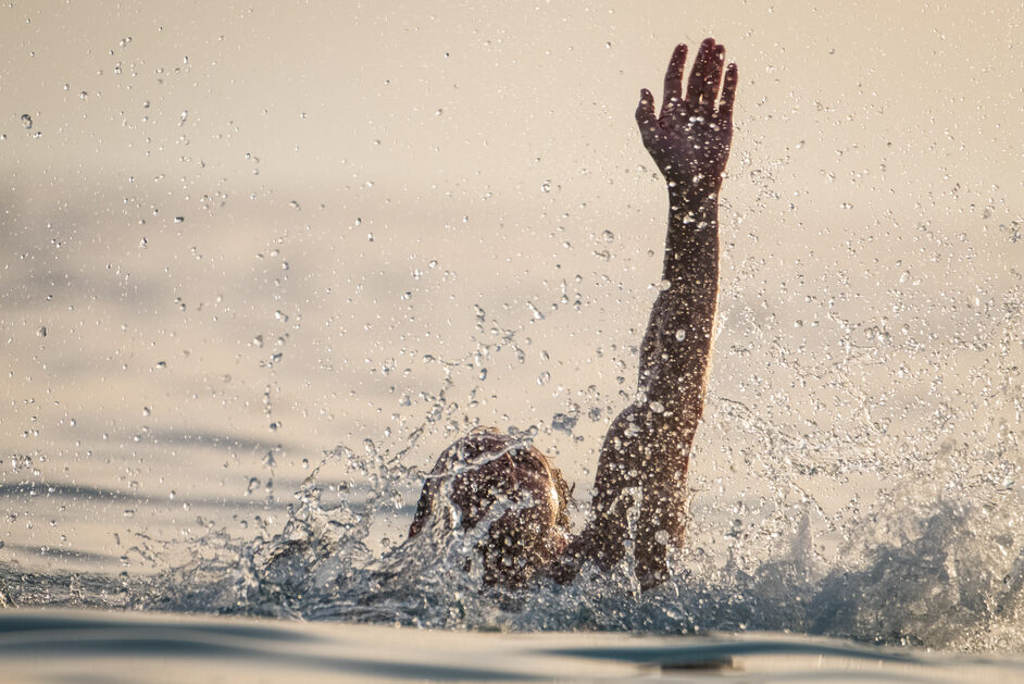 A person appears to be on the verge of drowning in the sea. One hand is raised, the head slightly above the surface, while the rest of the body is underwater.