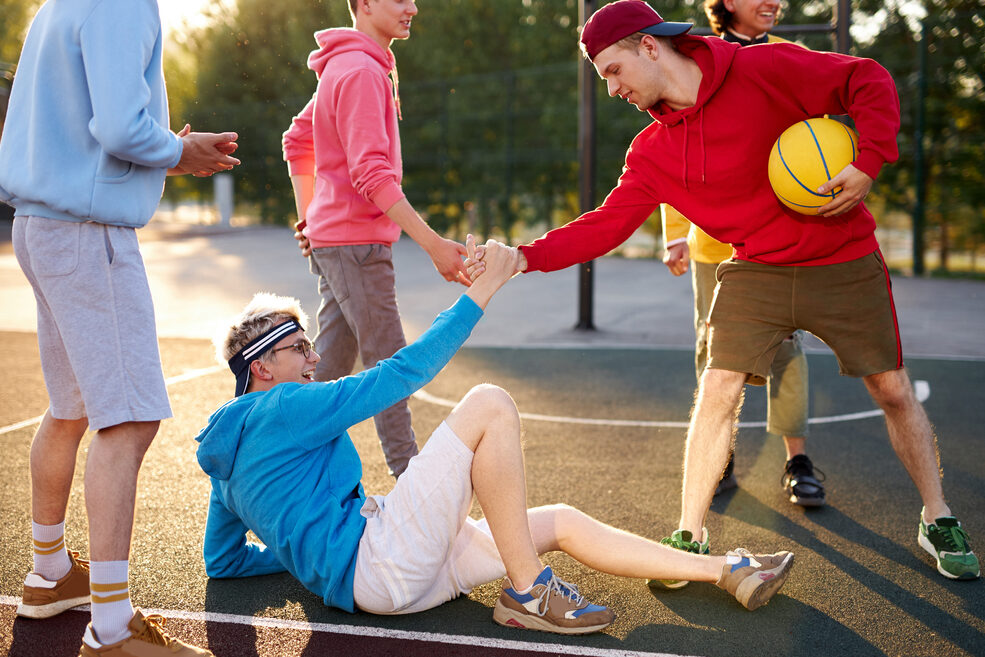 Young people are playing street basketball. One player has fallen, and another helps them up by offering a hand.