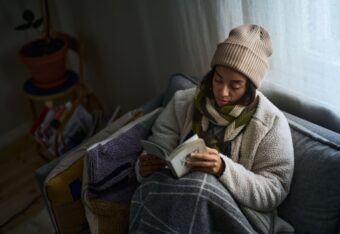 Nainen istuu sohvalla kirja kädessä takki, kaulahuivi ja pipo päällä.