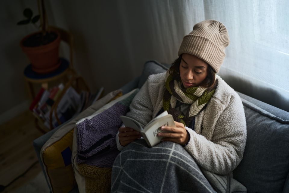 Nainen istuu sohvalla kirja kädessä takki, kaulahuivi ja pipo päällä.