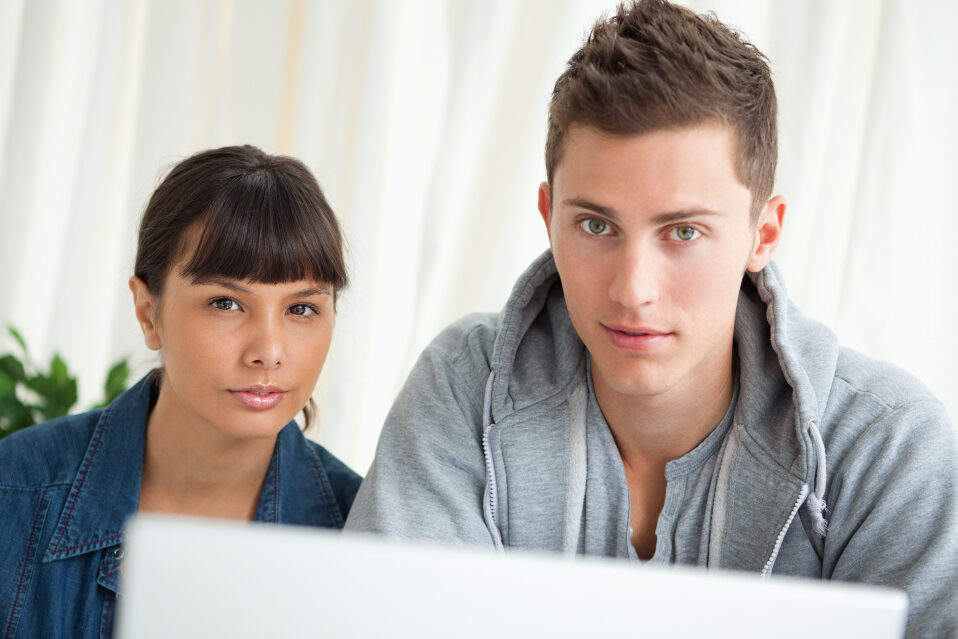 A young woman and man sit with serious expressions in front of a laptop.