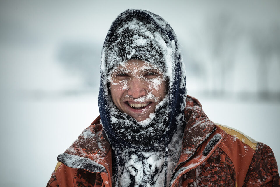 A smiling young man stands in a snowy landscape, his face covered with snow and dressed in winter clothing.
