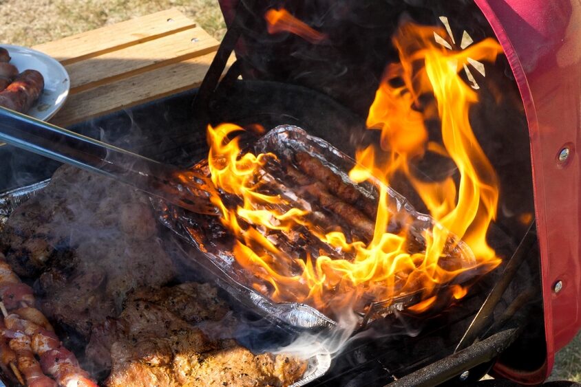 A disposable foil tray with sausages on fire inside a charcoal grill.