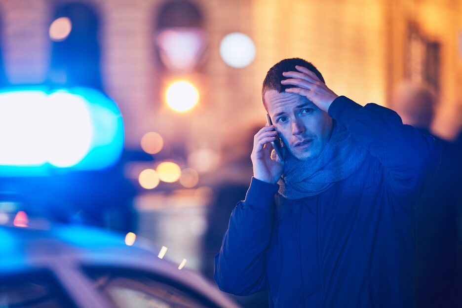 A young man standing on a street, talking on his phone and holding his head, looking worried. A blue emergency vehicle light is flashing nearby.