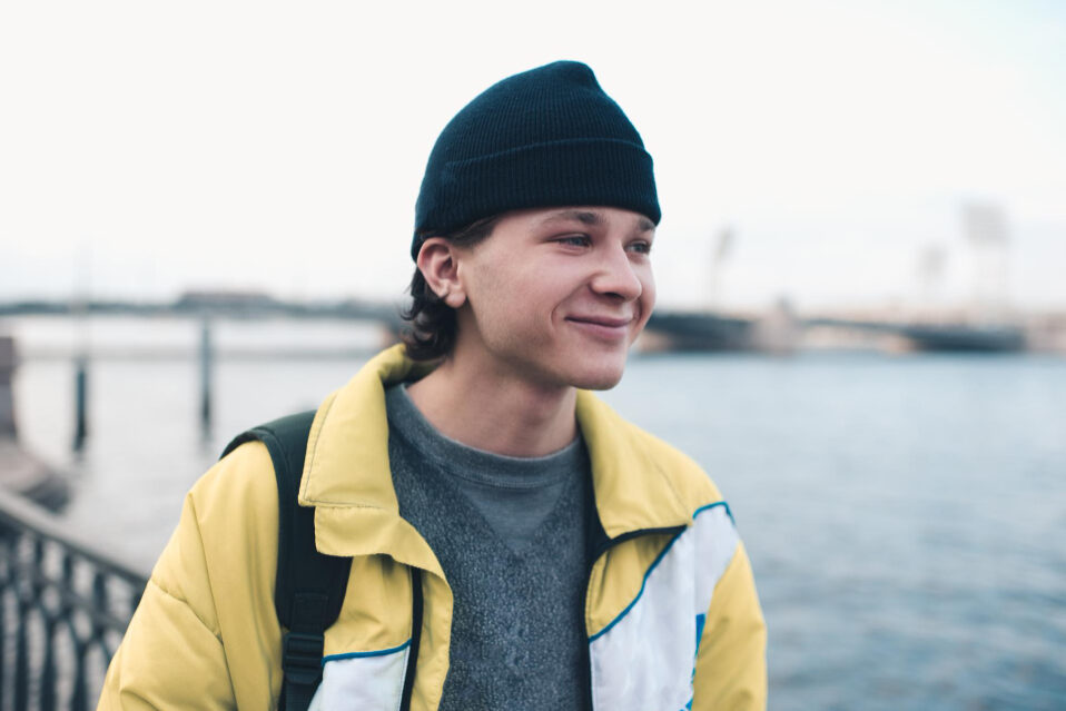 Two smiling young adults pose in firefighter gear, helmets under their arms, in front of a fire truck.