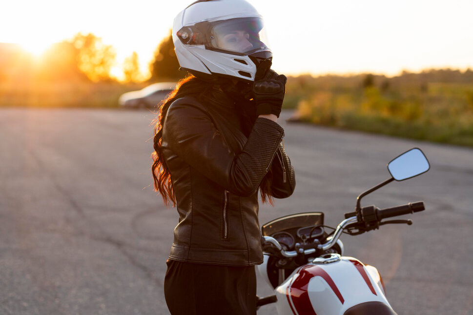 A motorcyclist stands on an asphalt lot with their bike, fastening their helmet.