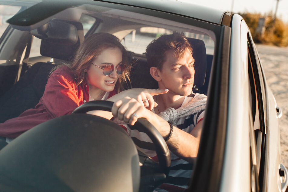 A young man and woman sit in a car on a sunny day, and the woman points at something outside the car.
