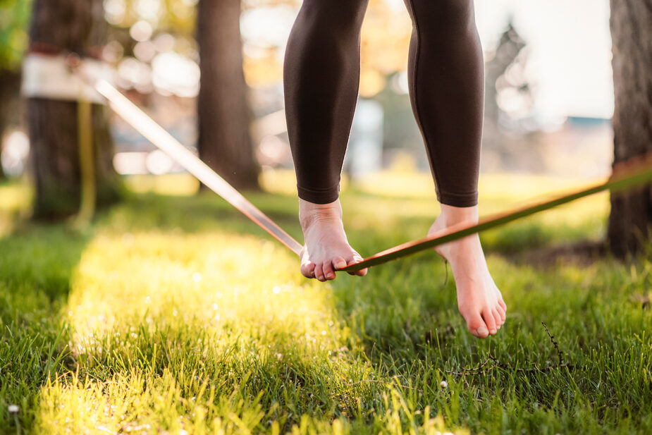 In a green park, a slackline is stretched between trees, and one person walks barefoot along it.