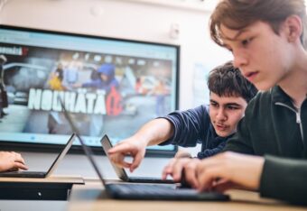 Kaksi teini-ikäistä poikaa tietokoneilla luokkahuoneessa. Toinen pojista osoittaa sormella toisen ruutua.