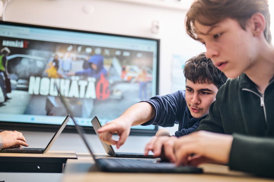 Kaksi teini-ikäistä poikaa tietokoneilla luokkahuoneessa. Toinen pojista osoittaa sormella toisen ruutua.