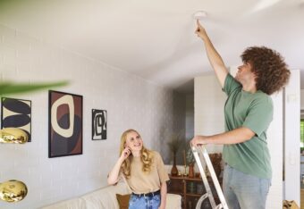 Nuori mies seisoo tikkailla olohuoneessa ja painaa palovaroittimen testinappia. Nuori nainen seuraa vierestä ja painaa toisella kädellä korvaa.