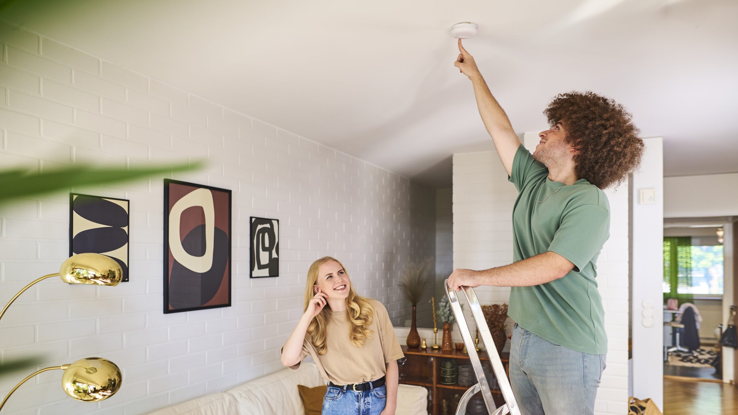 Nuori mies seisoo tikkailla olohuoneessa ja painaa palovaroittimen testinappia. Nuori nainen seuraa vierestä ja painaa toisella kädellä korvaa.