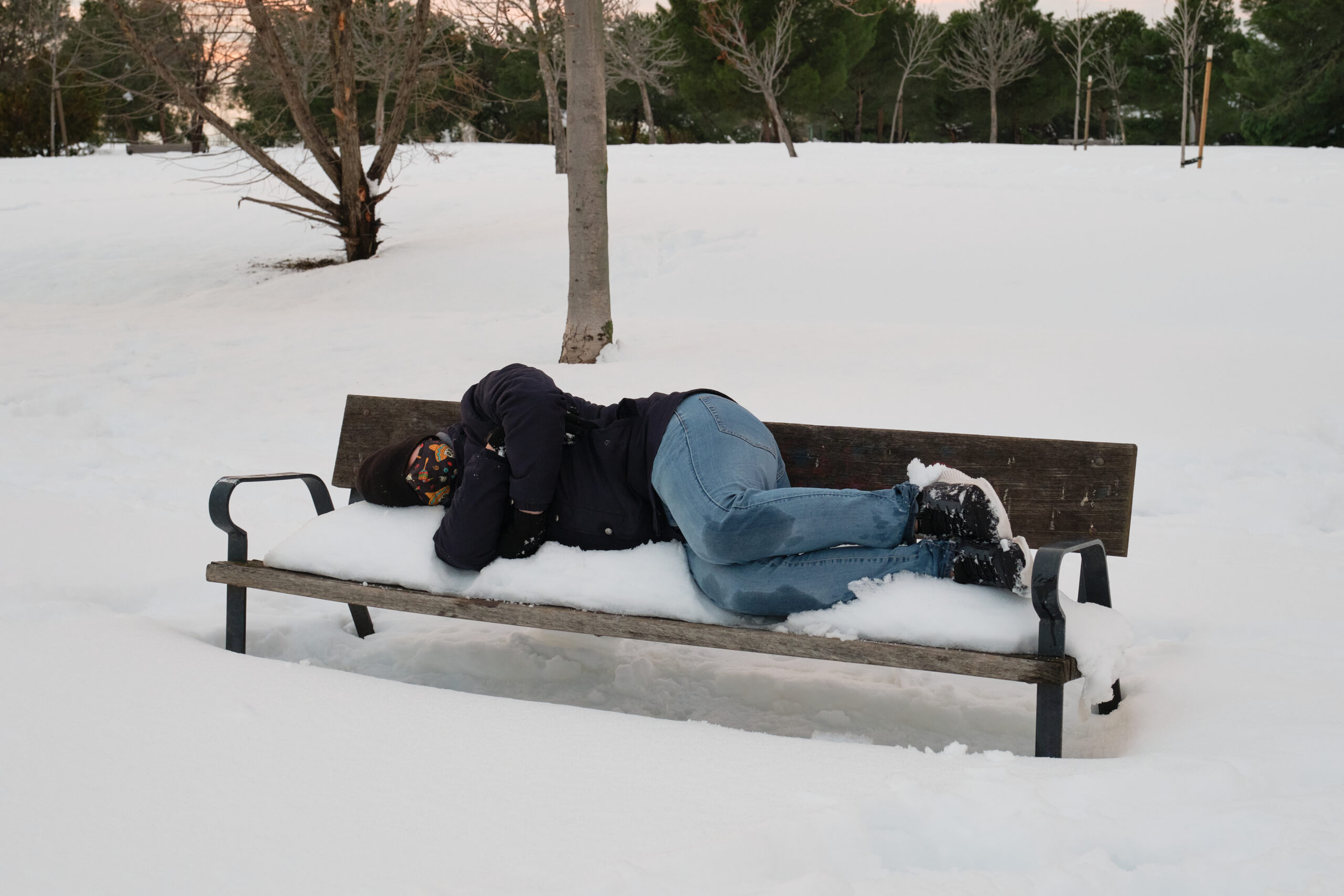 A person dressed in jeans, a thin dark jacket, and sneakers lies on a snowy park bench, appearing to be asleep.