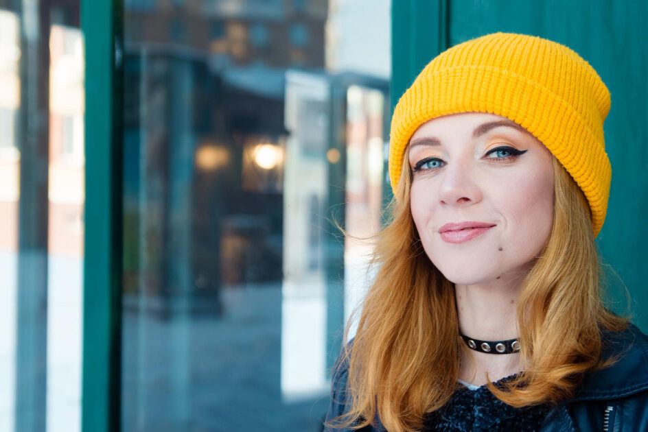 A young woman wearing a beanie stands next to a public transport stop, looking straight ahead with a determined, intense expression.