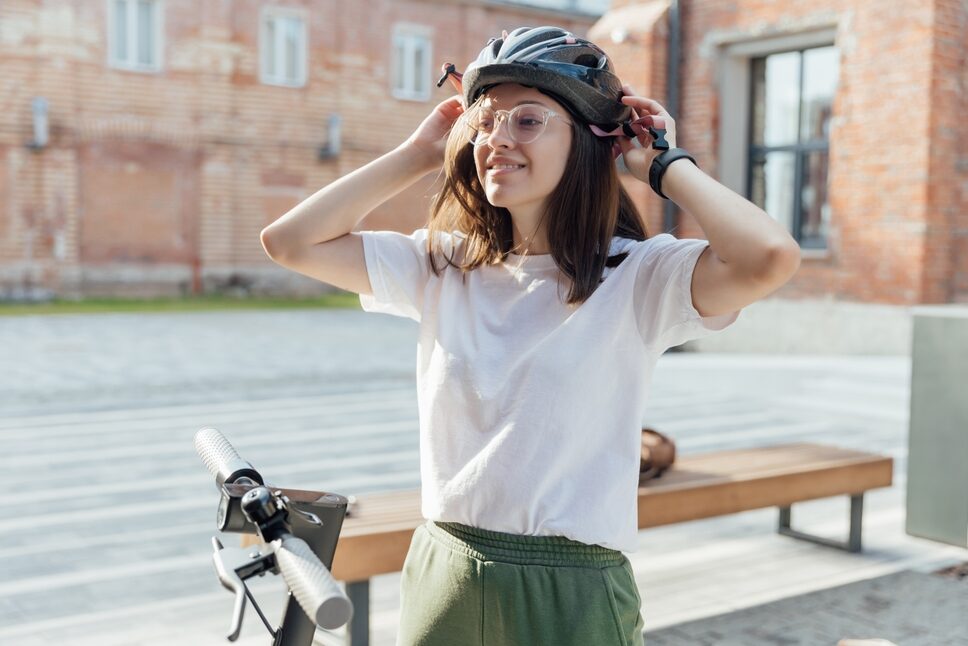 A smiling young woman stands on the street with her bicycle, adjusting her helmet.