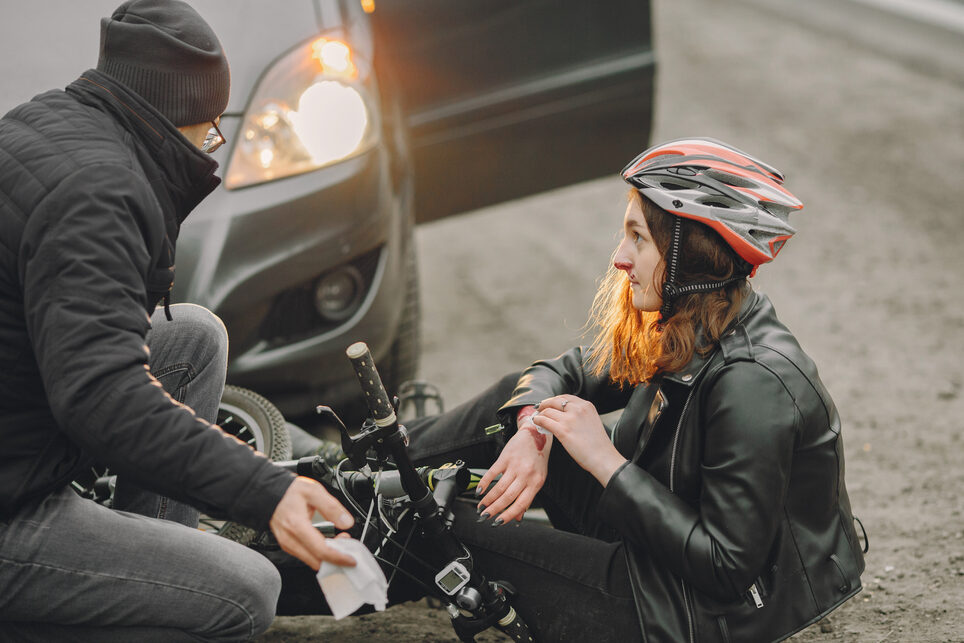 A woman with a bicycle helmet sits on the asphalt next to her bike, holding one wrist. She has a nosebleed. Beside her is a car, and someone has come out to help her.