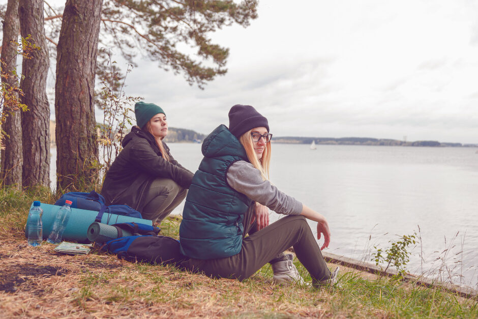 Two young women dressed in hiking gear sit in a forested area by the water.