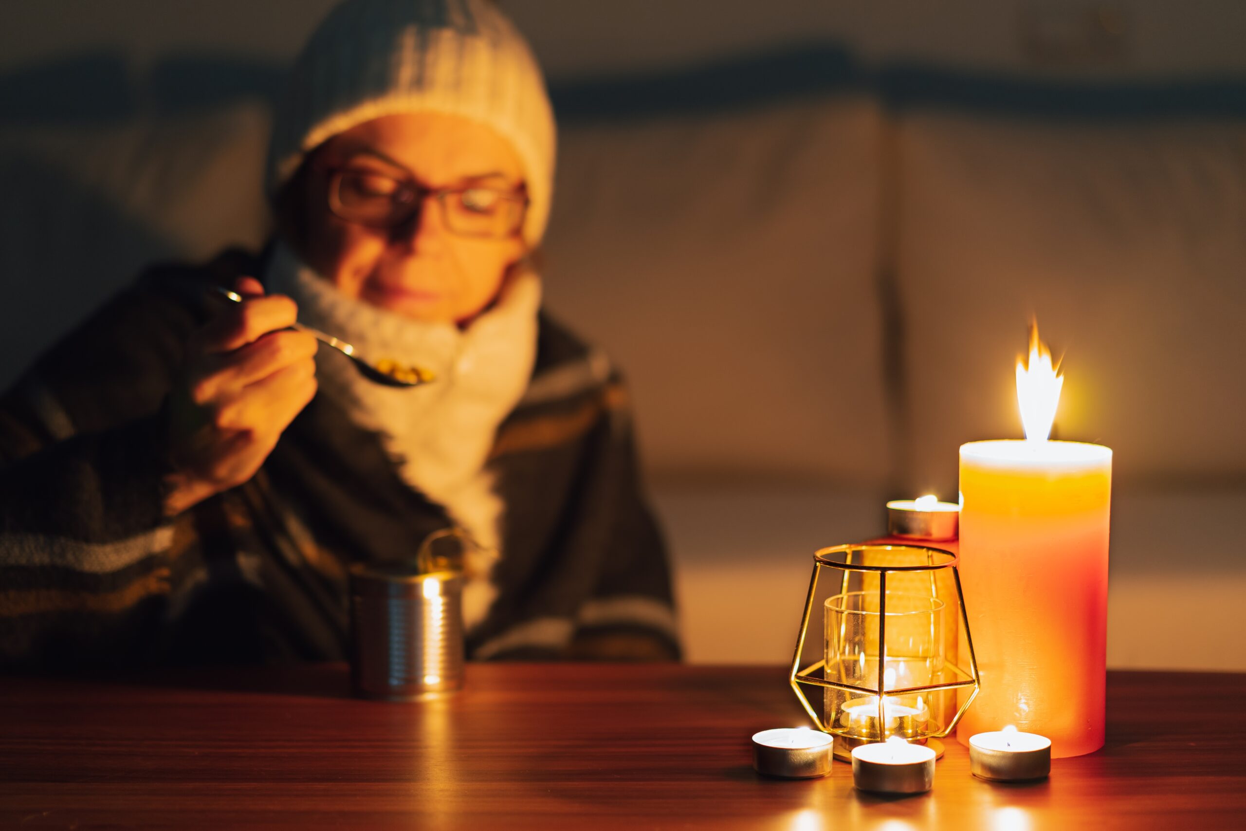 A young adult wearing a beanie and winter coat sits indoors by candlelight, eating food straight from a can.
