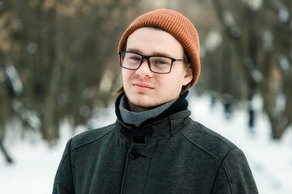 A young man wearing a beanie stands outdoors in a winter landscape, gazing thoughtfully into the distance.