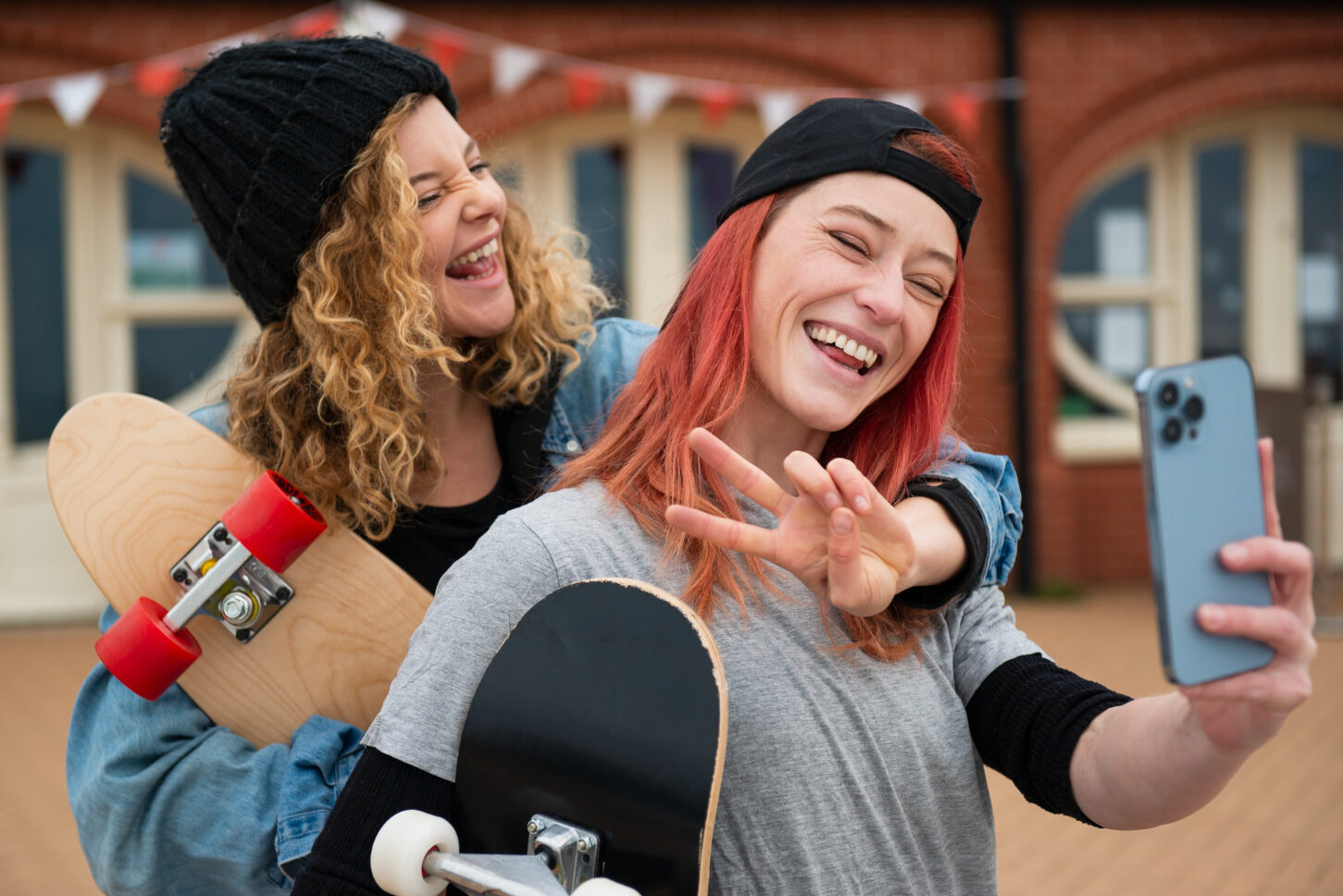 Two smiling young women pose for another woman’s phone. They hold skateboards, and one flashes a peace sign.