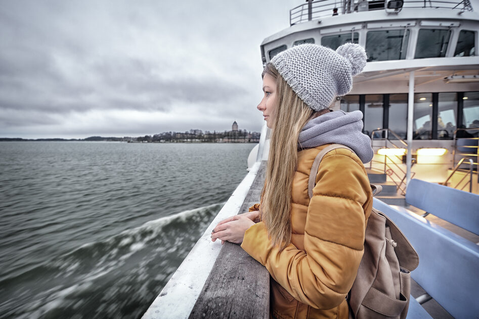A young woman wearing a beanie stands on the deck of the Suomenlinna ferry, looking out at the sea.