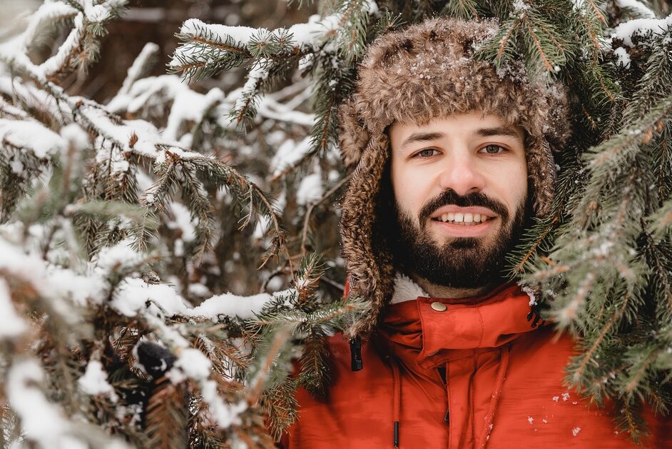 A dark-bearded man wearing a winter coat and fur hat stands among snowy spruces, smiling at the camera.
